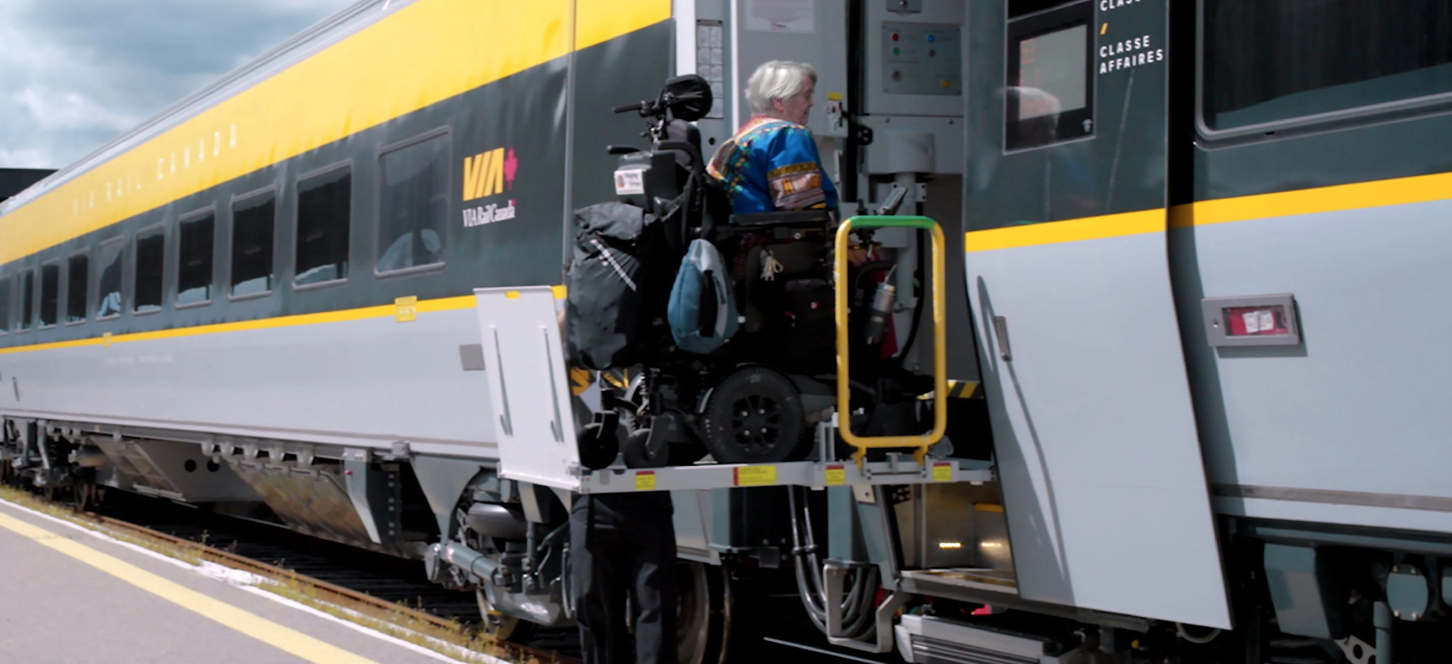 Une passagère en fauteuil roulant embarque à bord d'un train VIA Rail sur une plateforme élévatrice, depuis un quai extérieur.